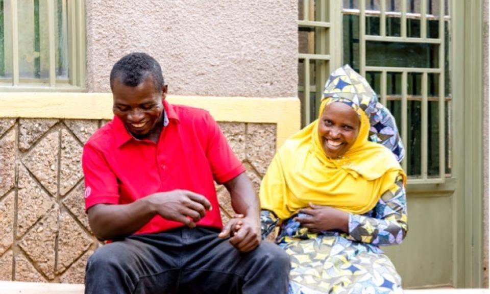 Two individuals sit on a bench outside a building with yellow walls and windows with metal grills. They are both laughing and holding hands.