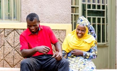 Two individuals sit on a bench outside a building with yellow walls and windows with metal grills. They are both laughing and holding hands.