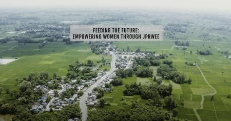 This image shows an aerial view of a rural landscape, primarily consisting of expansive green fields, likely rice paddies, interspersed with clusters of trees and small houses along a winding road. In the foreground, a small village or settlement is visible, with the road cutting through it. In the upper part of the image, there is a text overlay that reads: "FEEDING THE FUTURE: EMPOWERING WOMEN THROUGH JPRWEE."