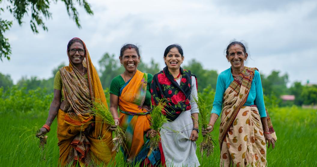 The image shows four smiling women standing in a field, all wearing brightly coloured saris.